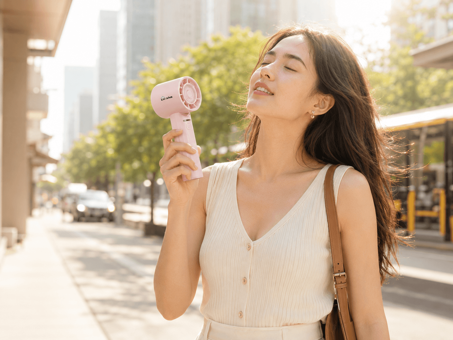 Woman with closed eyes uses a pink portable fan on a sunny city street.