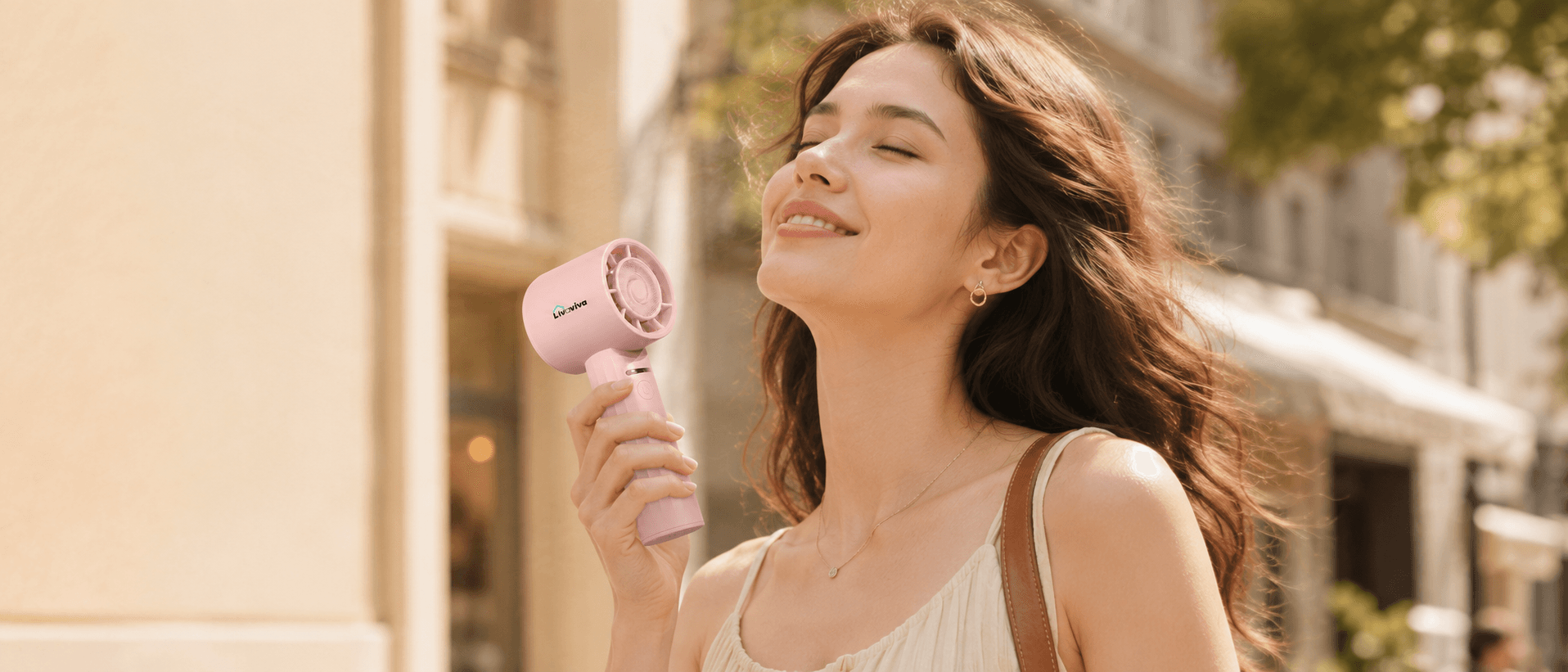 Smiling woman with closed eyes uses a pink handheld fan on a sunny street.